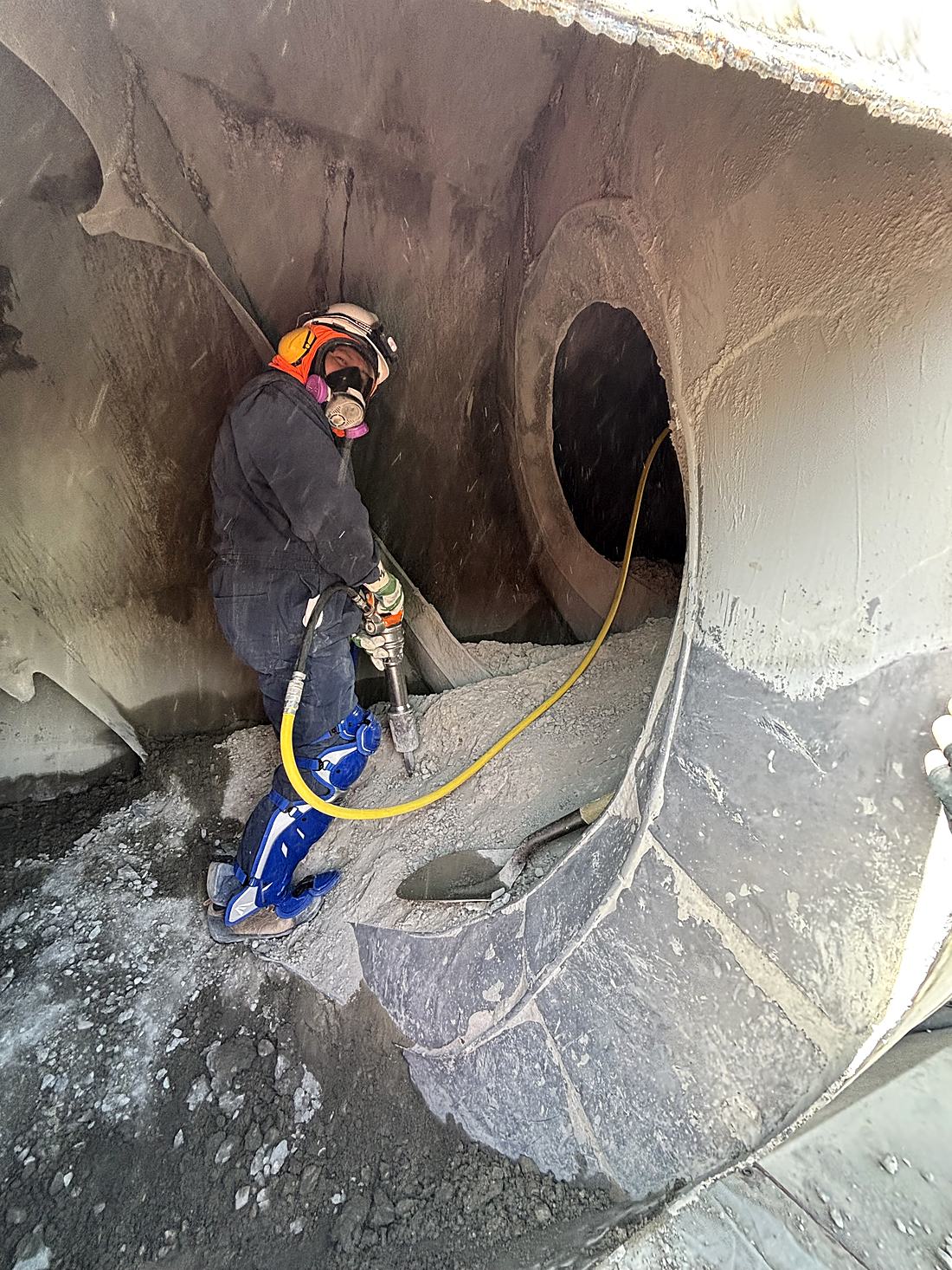 Express Chipping technician in respirator, hearing protection, and leg guards operating a pneumatic chipping hammer inside a ready mix drum